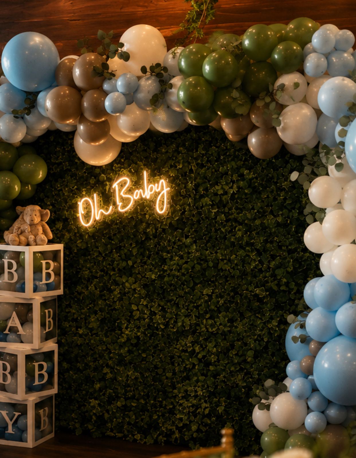 Baby shower backdrop at Kaskade — a balloon arch in blue, sage and gold against a boxwood wall with an Oh Baby neon sign