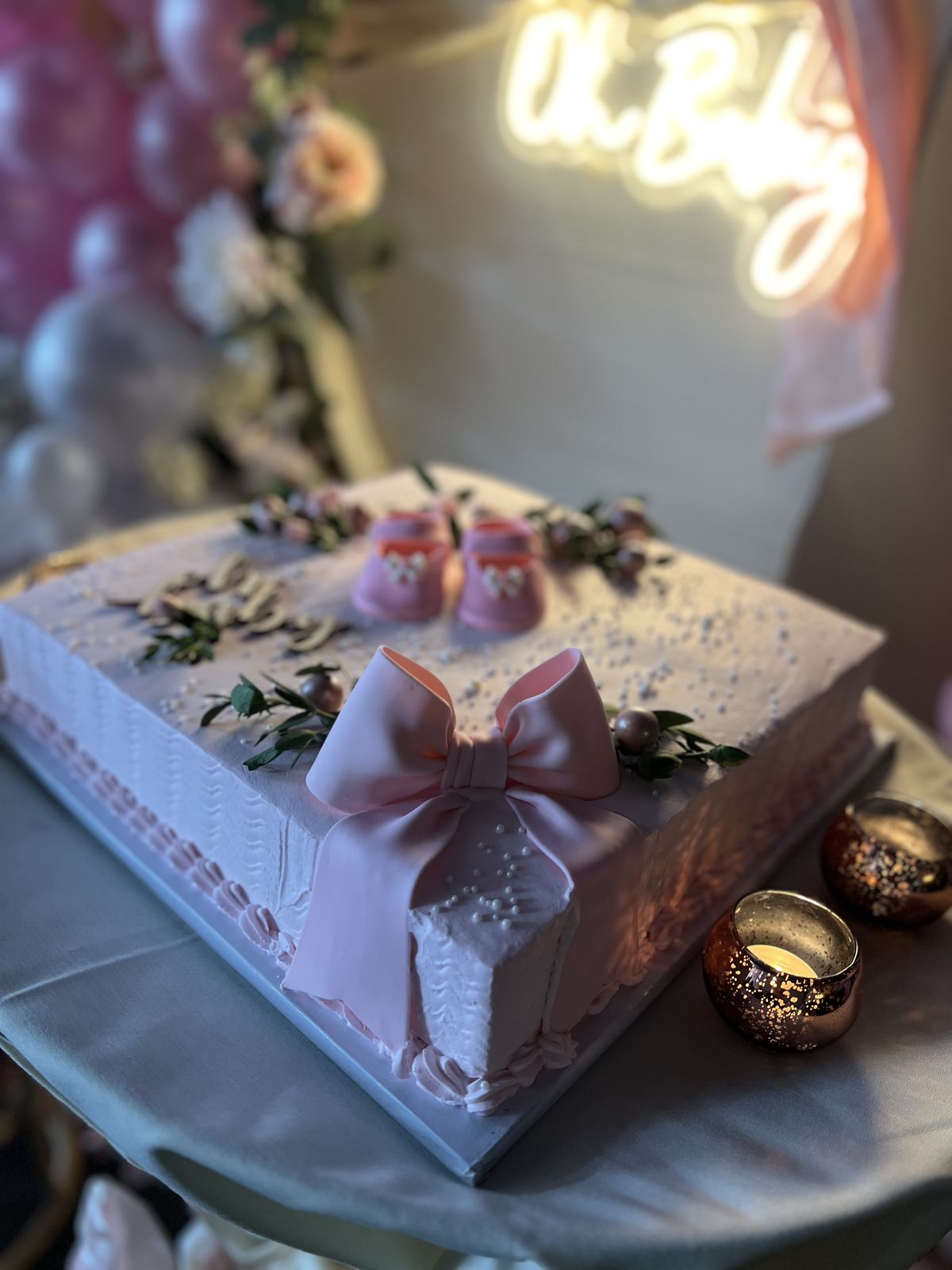 A pink and white celebration cake decorated with sugar flowers and baby shoes